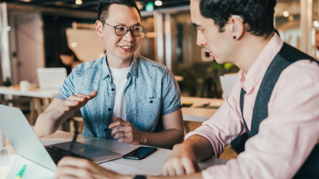 Two men talking with their hands at a table near laptop